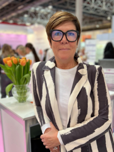 Lächelnde Frau mit großer Brille und breit gestreiftem schwarz-weißen Blazer lehnt an einem Verkaufstresen mit orangenem Tulpenstrauss links von ihr.